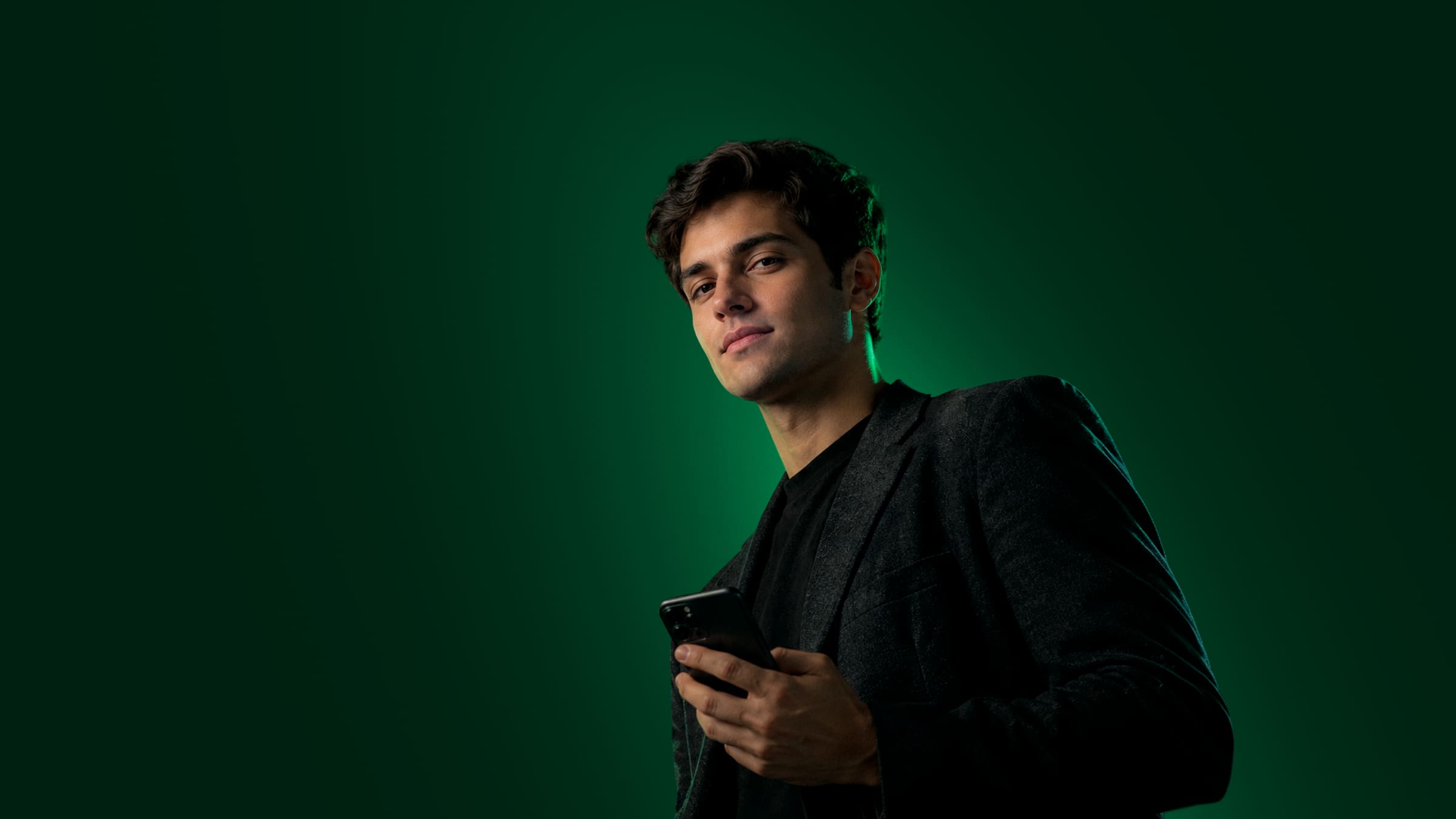 Homem de blazer escuro olhando o celular, iluminado por um brilho verde sobre fundo escuro.
